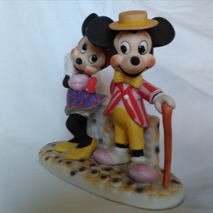 Może przedstawiać: Porcelanowa figurka Myszki Miki i Minnie Mouse. Miki ma na sobie czerwoną i białą w paski kurtkę, ż&oacute;łte spodnie i ż&oacute;łty kapelusz. Minnie ma na sobie fioletową i białą w kropki sukienkę. Stoją na podstawie, kt&oacute;ra wygląda jak brukowana ulica.