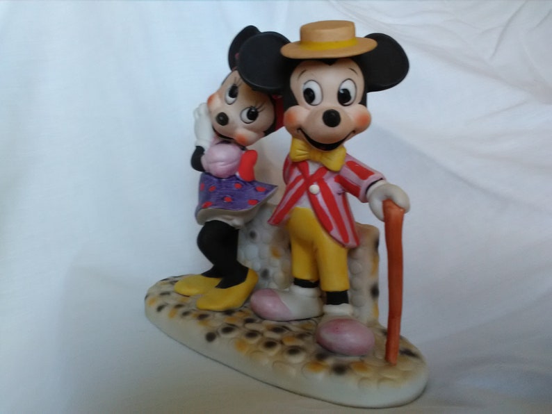 Może przedstawiać: Porcelanowa figurka Myszki Miki i Minnie Mouse. Miki ma na sobie czerwoną i białą w paski kurtkę, ż&oacute;łte spodnie i ż&oacute;łty kapelusz. Minnie ma na sobie fioletową i białą w kropki sukienkę. Stoją na podstawie, kt&oacute;ra wygląda jak brukowana ulica.