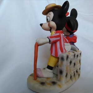 Może przedstawiać: Ceramiczna figurka przedstawiająca Myszki Miki i Minnie. Miki ma na sobie czerwoną i białą w paski koszulę, ż&oacute;łte spodnie i brązowy kapelusz. Trzyma laskę. Minnie ma na sobie fioletową sukienkę i czerwoną kokardkę. Stoją na białej podstawie z czarnymi kropkami.