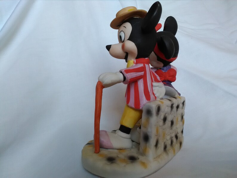 Może przedstawiać: Ceramiczna figurka przedstawiająca Myszki Miki i Minnie. Miki ma na sobie czerwoną i białą w paski koszulę, ż&oacute;łte spodnie i brązowy kapelusz. Trzyma laskę. Minnie ma na sobie fioletową sukienkę i czerwoną kokardkę. Stoją na białej podstawie z czarnymi kropkami.