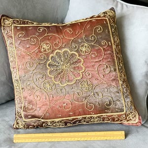 May include: A decorative throw pillow with a pink and brown velvet fabric and gold embroidery. The embroidery features a floral design with swirls and a large central flower.