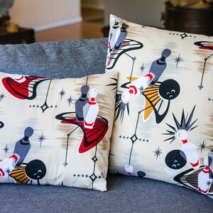 May include: Two decorative throw pillows with a retro bowling theme. The pillows feature a cream background with black, red, yellow, and white bowling pins and balls. The pillows have a star pattern and a mid-century modern design.