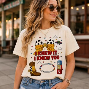 Pu&ograve; includere: Una t-shirt color crema con una stampa grafica di un fiocco giallo e bianco con motivo a macchie di mucca, uno stivale da cowboy marrone, un braccialetto di perline con il testo "YOU'VE GOT A FRIEND IN ME" e blocchi impilati con "TS 5" e "I KNEW IT I KNEW YOU."