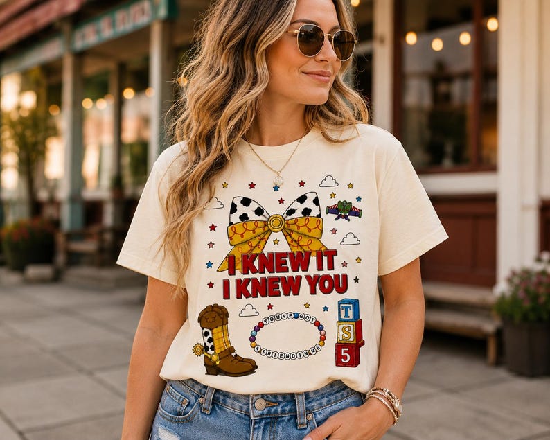 Pu&ograve; includere: Una t-shirt color crema con una stampa grafica di un fiocco giallo e bianco con motivo a macchie di mucca, uno stivale da cowboy marrone, un braccialetto di perline con il testo "YOU'VE GOT A FRIEND IN ME" e blocchi impilati con "TS 5" e "I KNEW IT I KNEW YOU."