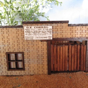 May include: A model of a wooden building with a sign that reads "O.K. Corral Feed and Livery Stables Horses Mules Bought Sold & Traded", a window, and a wooden gate.