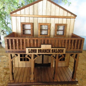 May include: A wooden model of a saloon with a balcony and a sign that reads "Long Branch Saloon". The saloon is made of light brown wood and has a dark brown roof. The balcony is made of dark brown wood and has a railing. The sign is made of dark brown wood and has white lettering.