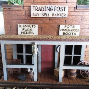 May include: A wooden model of a trading post with a sign that reads "Trading Post Buy Sell Barter". The building has a porch with two chairs and a lantern hanging from the ceiling. There are signs above the doors that read "Blankets Furs Hides" and "Pots Canned Goods Boots".