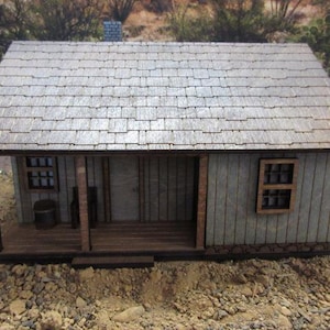 May include: A small wooden model of a house with a shingled roof, two windows, and a porch. The house is painted a light gray color and has a brown wooden door.