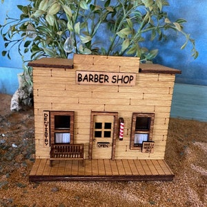 May include: A wooden model of a barber shop with a sign that reads "Barber Shop". The shop has a door with a barber pole and a sign that says "Open". There is a bench in front of the shop.