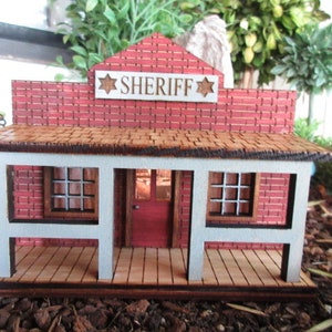 May include: A miniature wooden sheriff's office building with a red brick facade, a white porch, and a sign that reads "SHERIFF" with two stars.