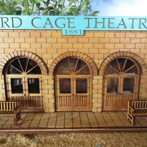 May include: A wooden model of a brick building with three arched doorways and a sign that reads "BIRD CAGE THEATRE 1881". The building has two wooden benches in front of it.