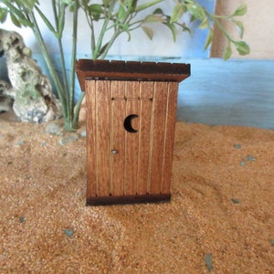 May include: A miniature wooden outhouse with a brown wooden door and a crescent moon cut-out. The outhouse is sitting on a sandy surface.