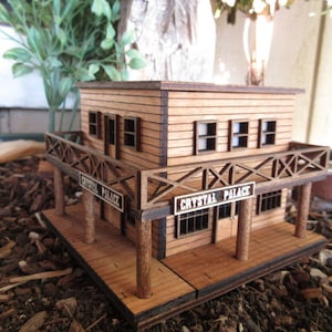 May include: A wooden model of a two-story building with a balcony and a sign that reads "Crystal Palace". The building is painted brown and has white trim. The model is sitting on a bed of brown wood chips.