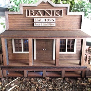May include: A wooden model of a Western-style bank building with a sign that reads "BANK Est. 1876 Assay & Land Offices".