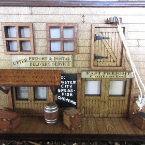 May include: A wooden model of a Western-style storefront with a sign that reads "Utter Freight & Postal Delivery Service" and "Fast Freight Contracted". The storefront has a wooden awning, two windows, and a door. There is a sign on the side of the building that reads "0: Custer City Spear-Fish Cheyenne".