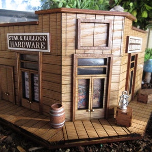 May include: A miniature wooden building with a sign that reads "Star & Bullock Hardware". The building has a brown wooden facade with a window and a door. There is a barrel in front of the building.