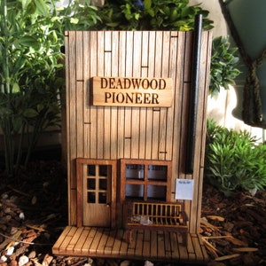 May include: A wooden model of a building with a sign that reads "Deadwood Pioneer". The building has a door and a window, and there is a bench in front of it.