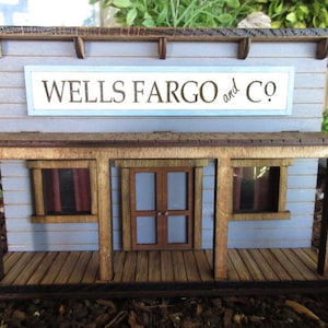 May include: A miniature wooden building with a sign that reads "WELLS FARGO and CO.". The building has a porch with two windows and a door. The building is painted a light blue color.