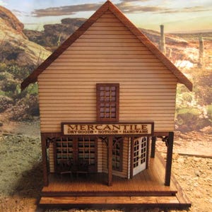 May include: A wooden model of a Western-style mercantile store with a sign that reads "Mercantile Dry Goods - Notions - Hardware". The store has a front porch with a wooden floor and a window on the front.