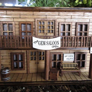 May include: A wooden model of a western saloon with a sign that reads "The Gem Saloon Open". The saloon has two floors, a balcony, and a wooden barrel in front. The model is painted in shades of brown and has a rustic look.