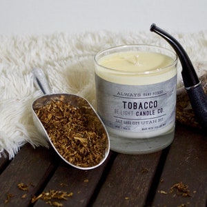 May include: A glass candle jar with a white candle inside. The label reads "Always Hand Picked Tobacco Be-Light Candle Co. Salt Lake City Utah 2012". There is a silver scoop filled with brown tobacco flakes, a black pipe, and a white fluffy rug in the background.