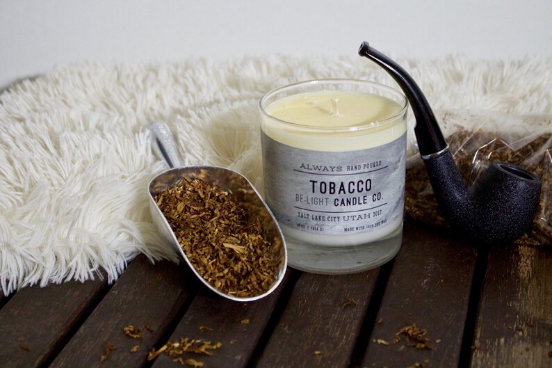 May include: A glass candle jar with a white candle inside. The label reads "Always Hand Picked Tobacco Be-Light Candle Co. Salt Lake City Utah 2012". There is a silver scoop filled with brown tobacco flakes, a black pipe, and a white fluffy rug in the background.