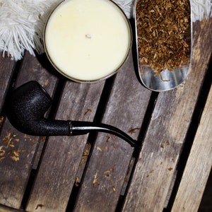 May include: A black pipe, a white candle, and a silver scoop filled with brown tobacco leaves on a wooden surface.