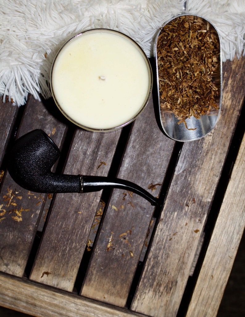 May include: A black pipe, a white candle, and a silver scoop filled with brown tobacco leaves on a wooden surface.