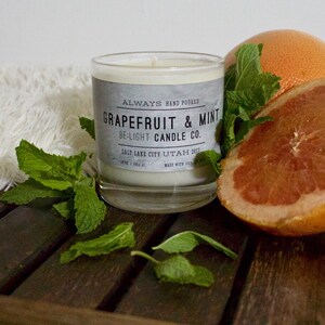 May include: A glass candle jar with a white label that reads "ALWAYS HAND POURED GRAPEFRUIT & MINT BE-LIGHT CANDLE CO. SALT LAKE CITY UTAH 2017". The candle is surrounded by fresh mint leaves and a halved grapefruit.