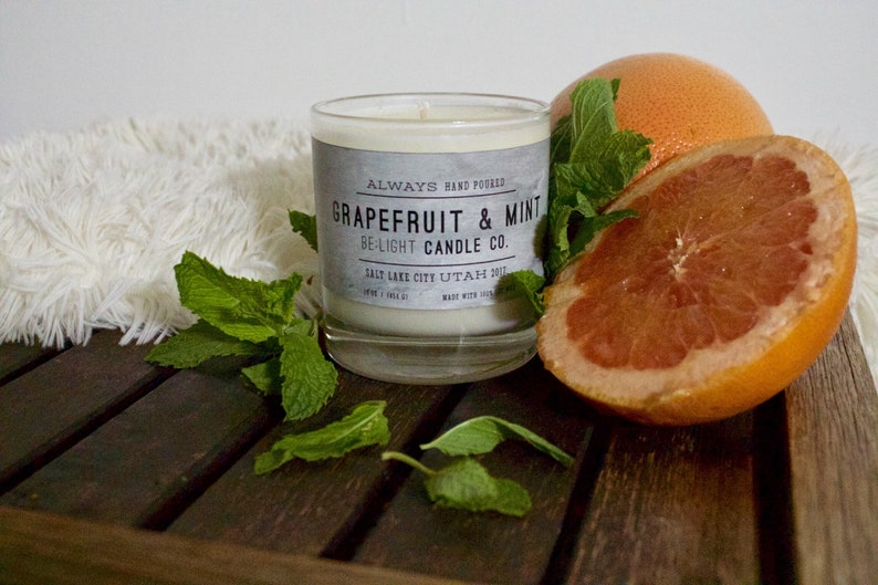 May include: A glass candle jar with a white label that reads "ALWAYS HAND POURED GRAPEFRUIT & MINT BE-LIGHT CANDLE CO. SALT LAKE CITY UTAH 2017". The candle is surrounded by fresh mint leaves and a halved grapefruit.
