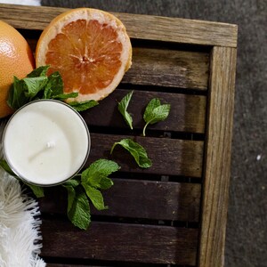 May include: A white candle with a wick in a glass jar, a halved grapefruit, and fresh green mint leaves on a wooden tray.