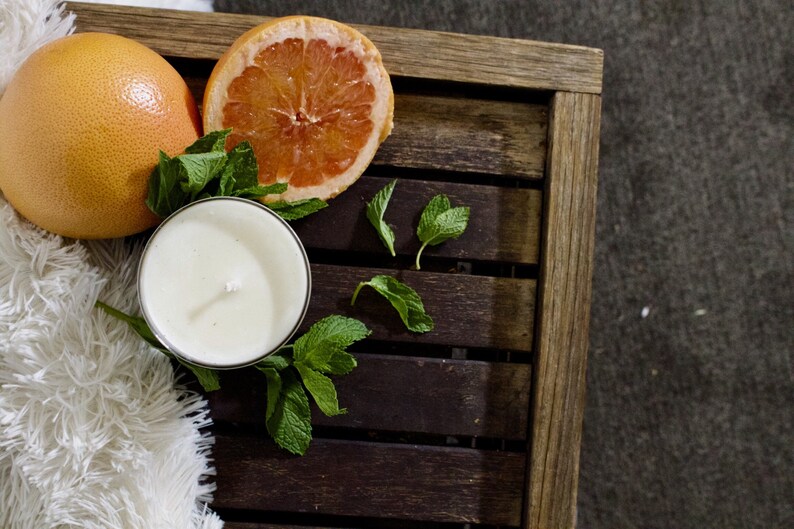 May include: A white candle with a wick in a glass jar, a halved grapefruit, and fresh green mint leaves on a wooden tray.