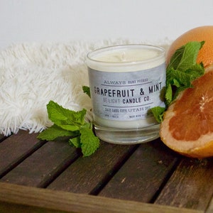 May include: A glass candle jar with a white label that reads "Always Hand Poured Grapefruit & Mint Be Light Candle Co. Salt Lake City, Utah 84111 Made with Soy Wax". The candle is surrounded by a grapefruit, a grapefruit slice, and fresh mint leaves on a wooden surface.