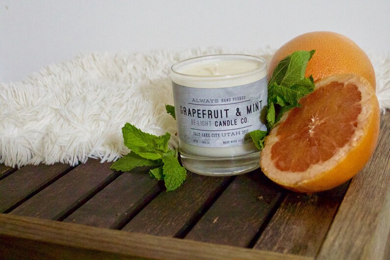 May include: A glass candle jar with a white label that reads "Always Hand Poured Grapefruit & Mint Be Light Candle Co. Salt Lake City, Utah 84111 Made with Soy Wax". The candle is surrounded by a grapefruit, a grapefruit slice, and fresh mint leaves on a wooden surface.