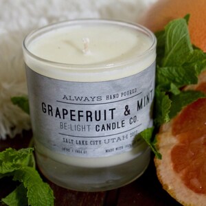 May include: A glass candle jar with a white label that reads "ALWAYS HAND POURED GRAPEFRUIT & MINT BE:LIGHT CANDLE CO. SALT LAKE CITY UTAH 2017". The candle is surrounded by fresh green mint leaves and a grapefruit slice.