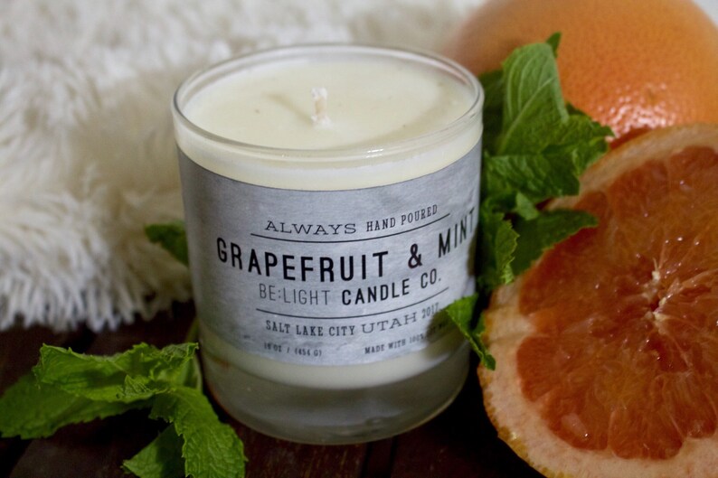 May include: A glass candle jar with a white label that reads "ALWAYS HAND POURED GRAPEFRUIT & MINT BE:LIGHT CANDLE CO. SALT LAKE CITY UTAH 2017". The candle is surrounded by fresh green mint leaves and a grapefruit slice.