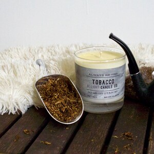 May include: A glass candle jar with a white candle inside. The jar has a label that reads "Always Hand Poured Tobacco Be-Light Candle Co. Salt Lake City, Utah 84112". There is a pipe and a scoop of tobacco leaves next to the candle.