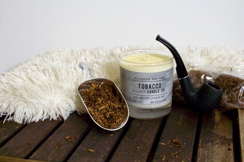 May include: A glass candle jar with a white candle inside. The jar has a label that reads "Always Hand Poured Tobacco Be-Light Candle Co. Salt Lake City, Utah 84112". There is a pipe and a scoop of tobacco leaves next to the candle.