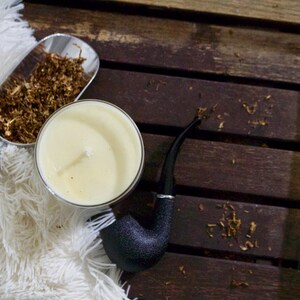 May include: A white candle in a glass jar, a silver scoop filled with brown tobacco, and a black pipe on a wooden surface.