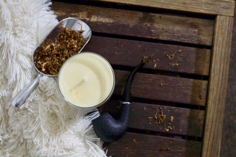 May include: A white candle in a glass jar, a silver scoop filled with brown tobacco, and a black pipe on a wooden surface.