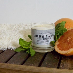 May include: A white candle in a glass jar with a black and white label that reads "ALWAYS HAND POURED Grapefruit & Mint BE-LIGHT CANDLE CO. SALT LAKE CITY UTAH 2021". The candle is surrounded by fresh mint leaves and a halved grapefruit.