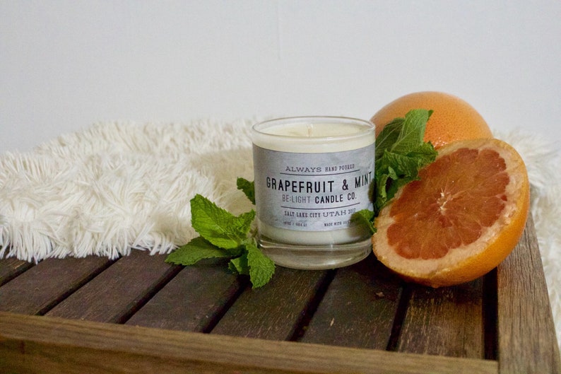 May include: A white candle in a glass jar with a black and white label that reads "ALWAYS HAND POURED Grapefruit & Mint BE-LIGHT CANDLE CO. SALT LAKE CITY UTAH 2021". The candle is surrounded by fresh mint leaves and a halved grapefruit.