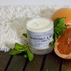 May include: A white candle with a gray label that reads "Always Hand Poured Grapefruit & Mint De-Light Candle Co. Salt Lake City Utah 84103 Made with Soy Wax"  The candle is surrounded by fresh green mint leaves and a halved grapefruit.