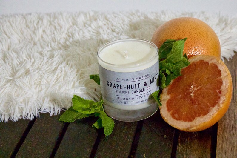 May include: A white candle with a gray label that reads "Always Hand Poured Grapefruit & Mint De-Light Candle Co. Salt Lake City Utah 84103 Made with Soy Wax"  The candle is surrounded by fresh green mint leaves and a halved grapefruit.