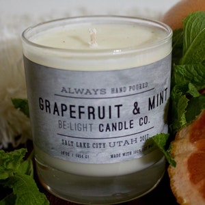 May include: A glass candle jar with a white label that reads "ALWAYS HAND POURED GRAPEFRUIT & MINT BE:LIGHT CANDLE CO. SALT LAKE CITY UTAH 2011. 18 oz / 1454 g MADE WITH 100% SOY WAX". The candle is surrounded by fresh green mint leaves and a grapefruit slice.