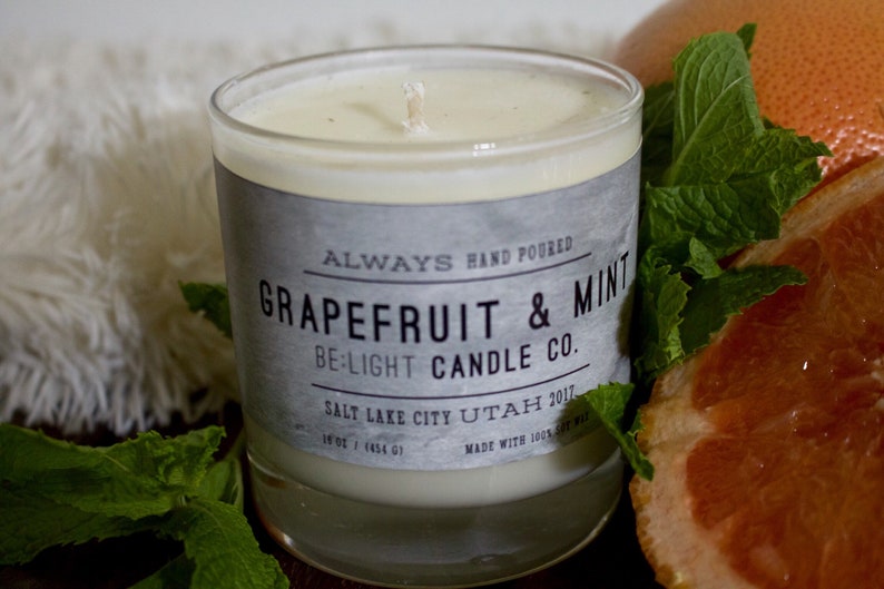 May include: A glass candle jar with a white label that reads "ALWAYS HAND POURED GRAPEFRUIT & MINT BE:LIGHT CANDLE CO. SALT LAKE CITY UTAH 2011. 18 oz / 1454 g MADE WITH 100% SOY WAX". The candle is surrounded by fresh green mint leaves and a grapefruit slice.
