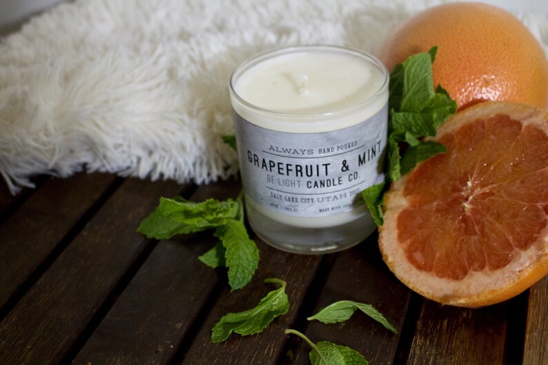 May include: A white candle in a glass jar with a gray label that reads "ALWAYS HAND POURED GRAPEFRUIT & MINT BE-LIGHT CANDLE CO. SALT LAKE CITY UTAH 84108 MADE WITH LOVE". The candle is surrounded by fresh green mint leaves and a grapefruit slice.