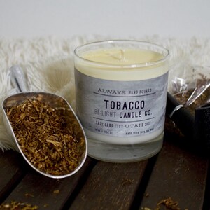 May include: A glass candle jar with a gray label that reads "ALWAYS HAND POURED TOBACCO BE-LIGHT CANDLE CO. SALT LAKE CITY UTAH 2017 MADE WITH 100% SOY WAX". The candle is surrounded by tobacco leaves and a pipe.