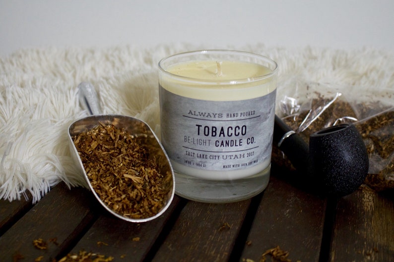 May include: A glass candle jar with a gray label that reads "ALWAYS HAND POURED TOBACCO BE-LIGHT CANDLE CO. SALT LAKE CITY UTAH 2017 MADE WITH 100% SOY WAX". The candle is surrounded by tobacco leaves and a pipe.