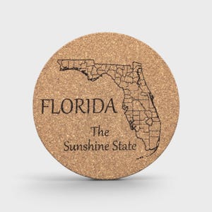 Puede incluir: Un posavasos redondo de corcho con un mapa de Florida grabado a láser en negro que muestra los límites de los condados. El texto FLORIDA aparece en una fuente serif negrita sobre el texto The Sunshine State en letra cursiva.
